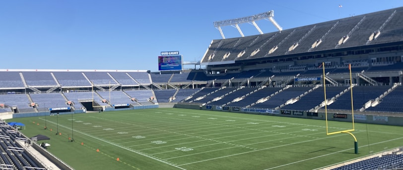 A large, empty sports stadium with blue seats surrounding a well-maintained green grass field. Bright sunlight creates clear shadows, and there are two goal posts on opposite ends of the field. A big electronic scoreboard displays some graphics, and the sky is clear and blue.